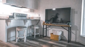 A compact kitchen with a built-in microwave, two stools, a wooden table with a flat-screen TV, and a wicker basket.