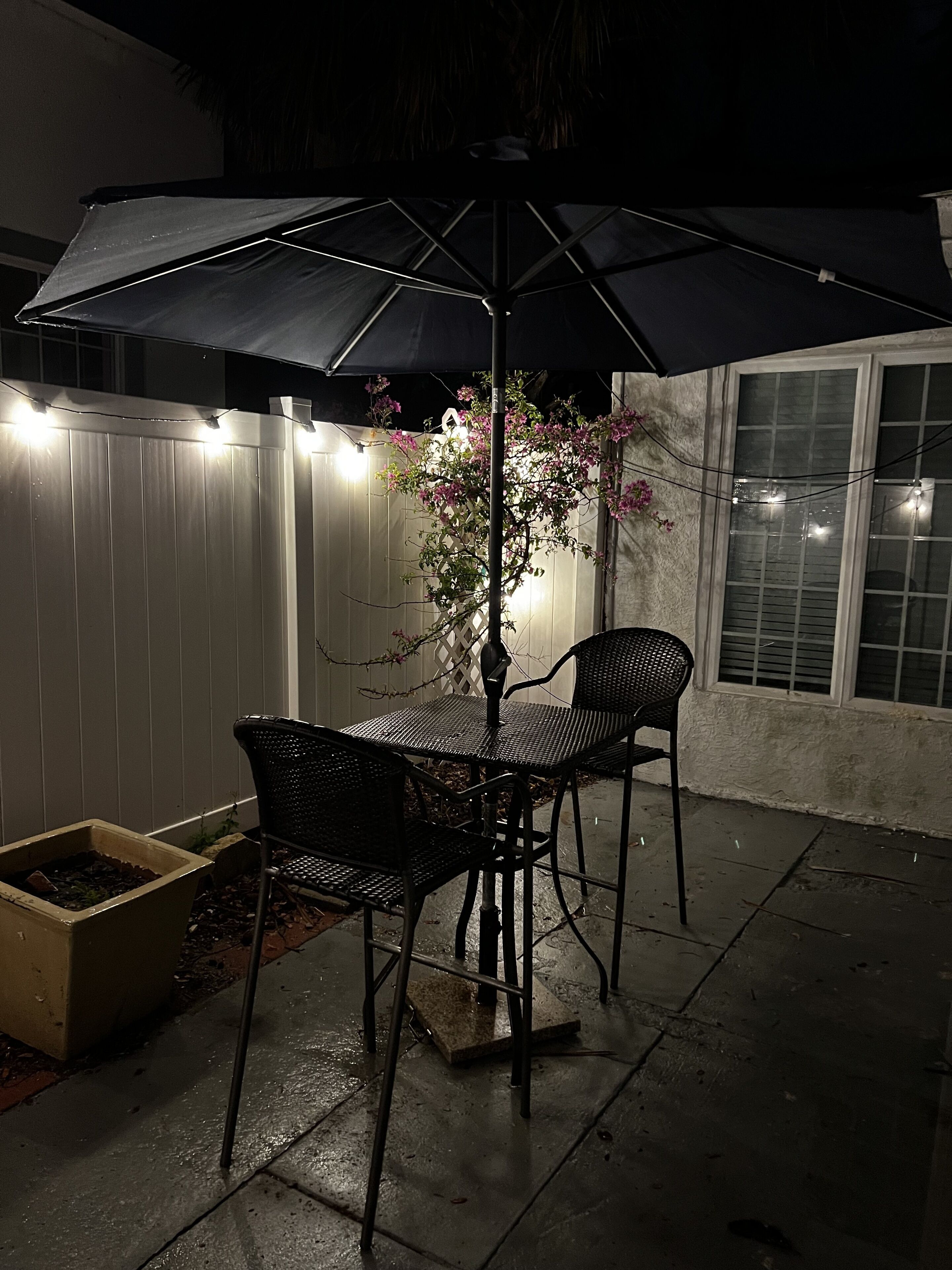 Une terrasse extérieure avec une table et des chaises sous un grand parasol, entourée d’une clôture blanche et illuminée de guirlandes lumineuses.