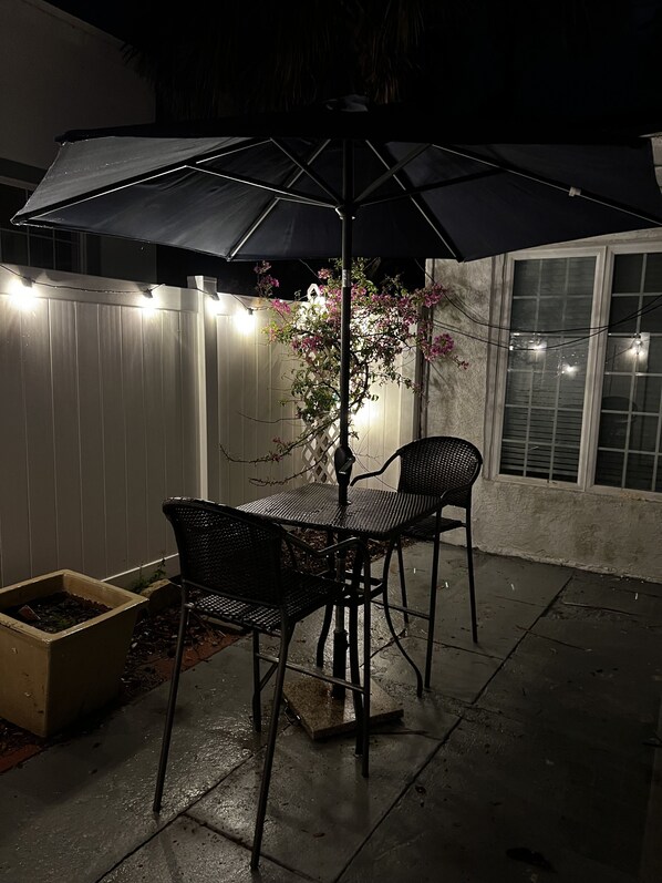 Une terrasse extérieure avec une table et des chaises sous un grand parasol, entourée d’une clôture blanche et illuminée de guirlandes lumineuses.