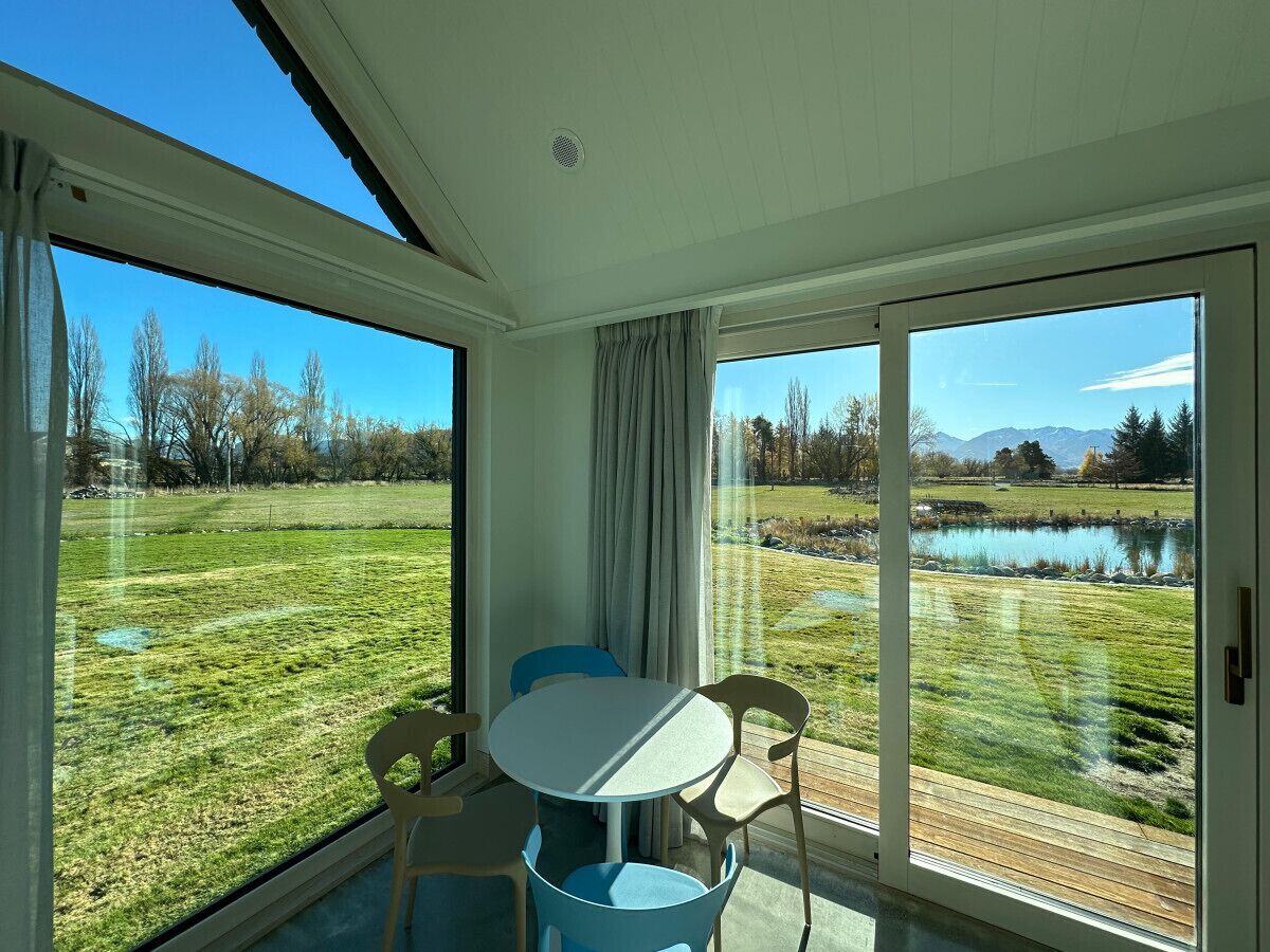 Une pièce avec une table et des chaises, de grandes fenêtres donnant sur un champ verdoyant et une étendue d’eau.