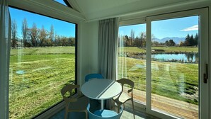 Une pièce avec une table et des chaises, de grandes fenêtres donnant sur un champ verdoyant et une étendue d’eau.