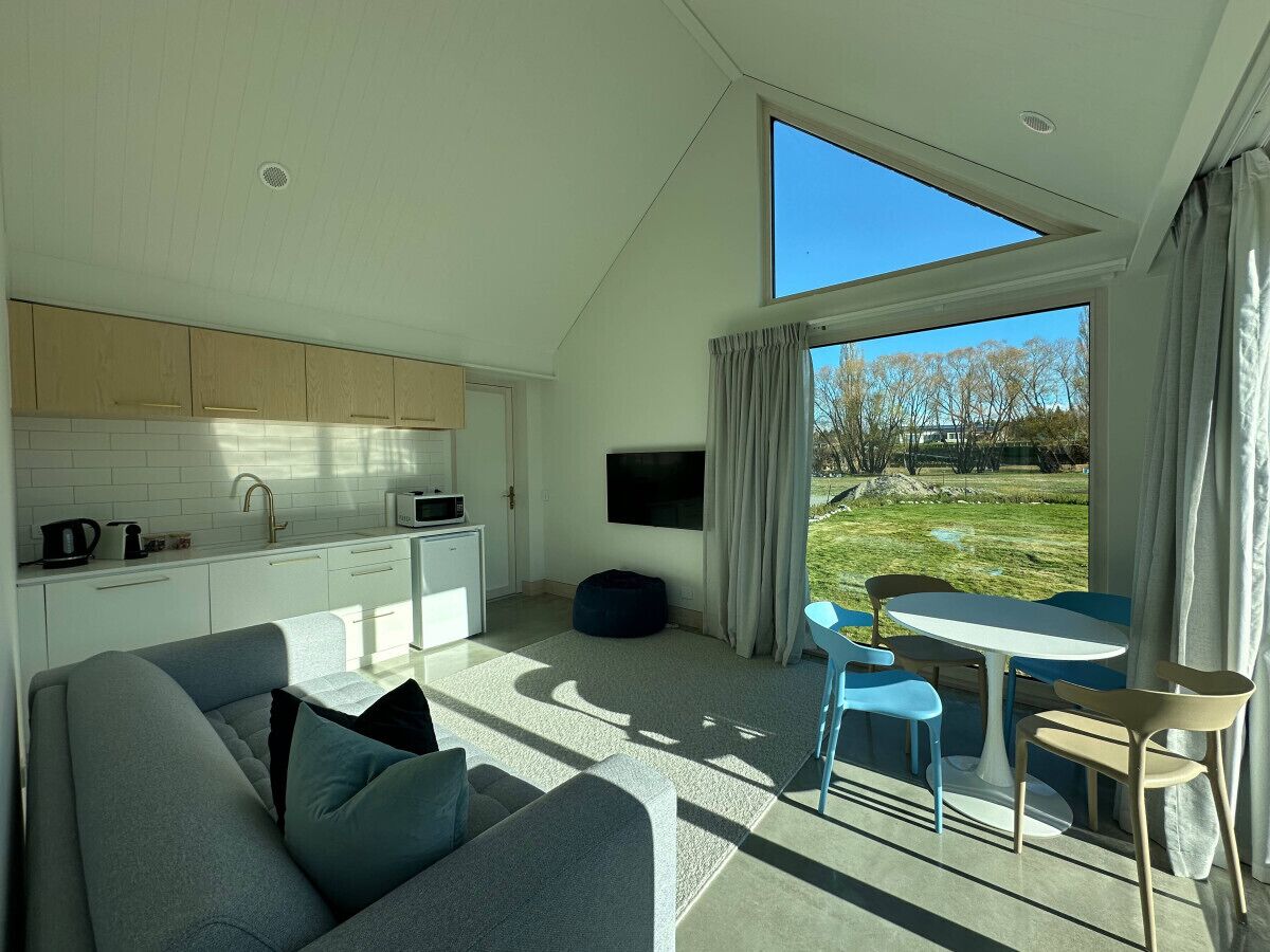 A modern living room with a sofa, a dining table with chairs, a kitchen area, and a large window with a view of the outdoors.