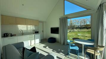 A modern living room with a sofa, a dining table with chairs, a kitchen area, and a large window with a view of the outdoors.