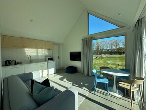 A modern living room with a sofa and a dining area with chairs and a table. There is a kitchen area with cabinets and a sink. A large window provides a view of the outdoors.