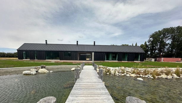 Une jetée en bois mène à un bâtiment moderne doté d’un toit sombre, et l’ensemble est entouré d’eau et de rochers.