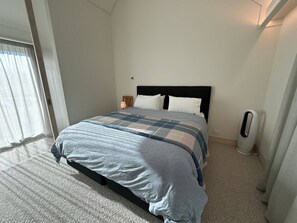 A neatly made bed with a checkered bedspread, a bedside table with a lamp, and a standing heater.