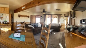 Interior acogedor de una cabaña con paredes de madera, comedor con mesa y sillas, cocina con microondas y tostadora, y sala de estar con sofá y chimenea.