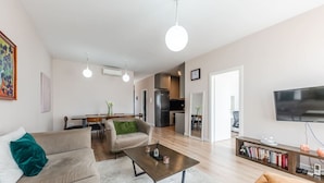 A modern living room with a sofa, coffee table, and a bookshelf. There is a dining area with chairs and a table, and a kitchen area with a refrigerator and stove. - 2-BR Apartment close to Radio Shkodra: Roger That! by PikHost (Shkodër)