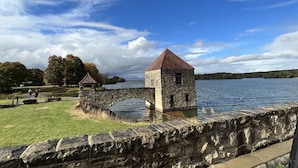 A stone structure with a red roof by a body of water, surrounded by a stone wall and grassy area. - Quaint Cabin #3 - Pymatuning Lake (Jamestown)