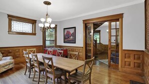 A dining area with a wooden table and chairs, a chandelier, a window with a stained glass design, and a door leading to another room. - Woodruff Manor 11 Bedroom Estate - 20+ guests (Indianapolis)