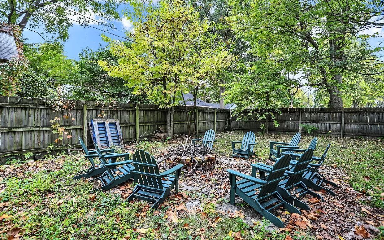 A backyard with a wooden fence, a fire pit, and several Adirondack chairs.