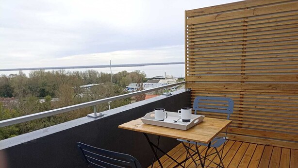 Ein Balkon mit Holztisch und zwei Stühlen, mit Blick auf ein Gewässer und entfernte Gebäude.