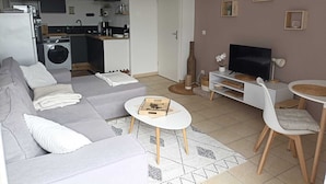 Ein modernes Wohnzimmer mit einem grauen Sofa, einem kleinen weißen Couchtisch, einem Fernseher und einem Essbereich mit weißem Tisch und Stühlen.