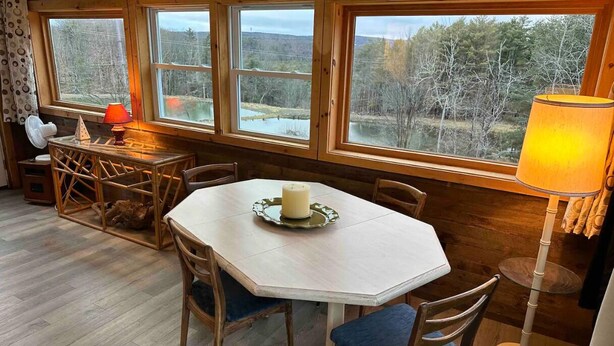 A cozy dining area with a round table, chairs, and a candle centerpiece. Large windows offer views of a forested landscape. - Magic Forest Farm's Artist Retreat (Coeymans Hollow)