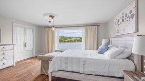 Ein Schlafzimmer mit Holzboden, einem Bett mit weißer Bettwäsche, einem Nachttisch, einer Kommode und einem großen Fenster mit Vorhängen.
