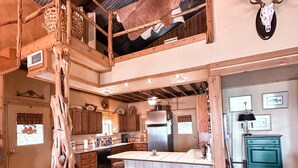 A rustic kitchen with a central island, wooden stools, and a high ceiling with exposed beams.