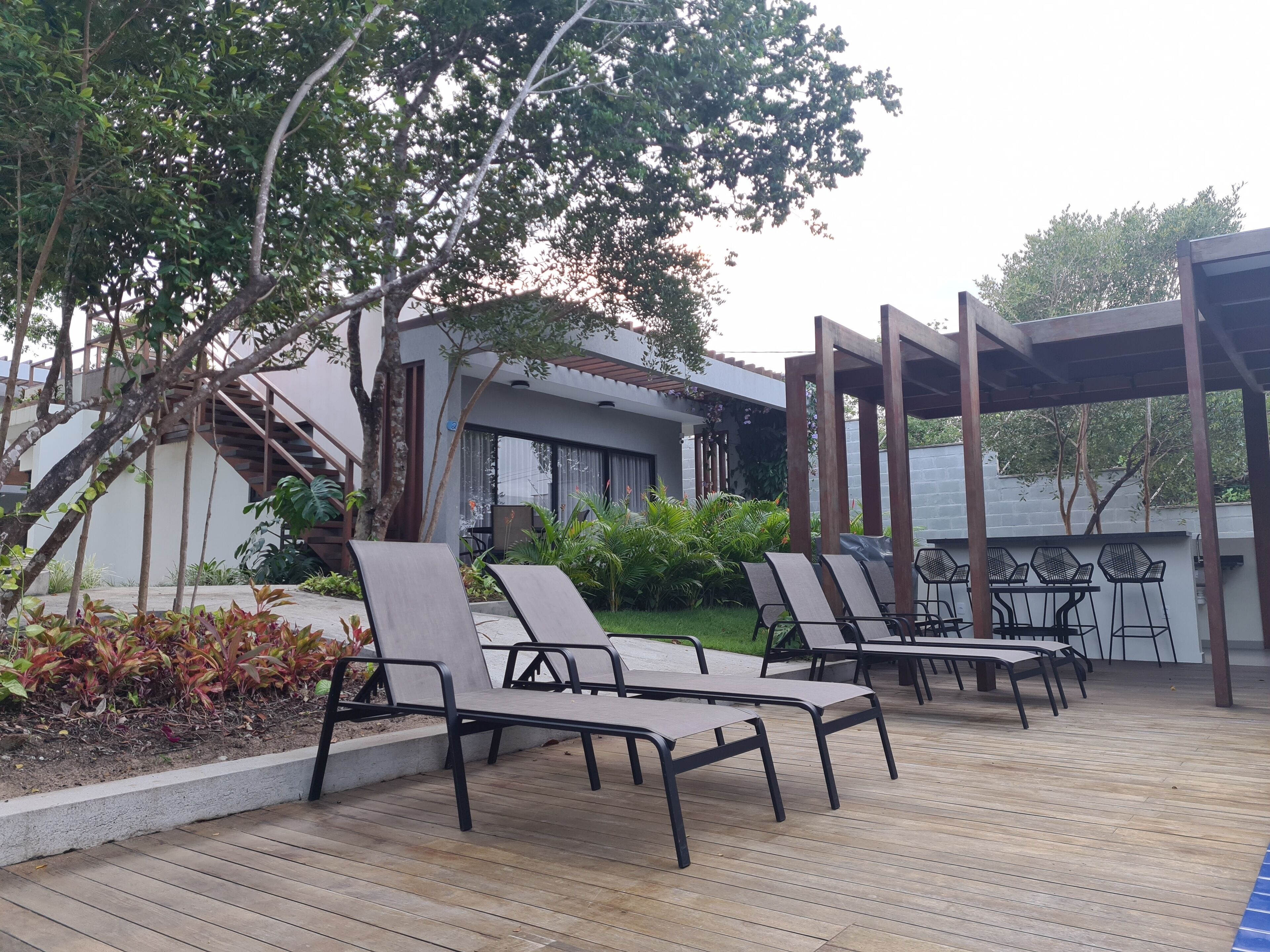 Une terrasse en bois avec des chaises longues et un espace abrité, entourée de verdure et bordée d’un bâtiment moderne doté d’un escalier.