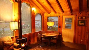 A cozy cabin interior with a wood-burning stove, a dining table with chairs, a sofa with a cushion, and a large window offering a view of the outside.