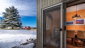 A snow-covered deck with a sliding glass door leading to a dining area with a patterned rug and wooden furniture. - Backwater Retreat Townhome Walk to Ski Shuttle (Driggs)
