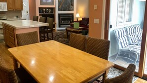 A dining area with a wooden table and chairs, a kitchen in the background, and a view of a balcony with striped outdoor furniture. - Backwater Retreat Townhome Walk to Ski Shuttle (Driggs)