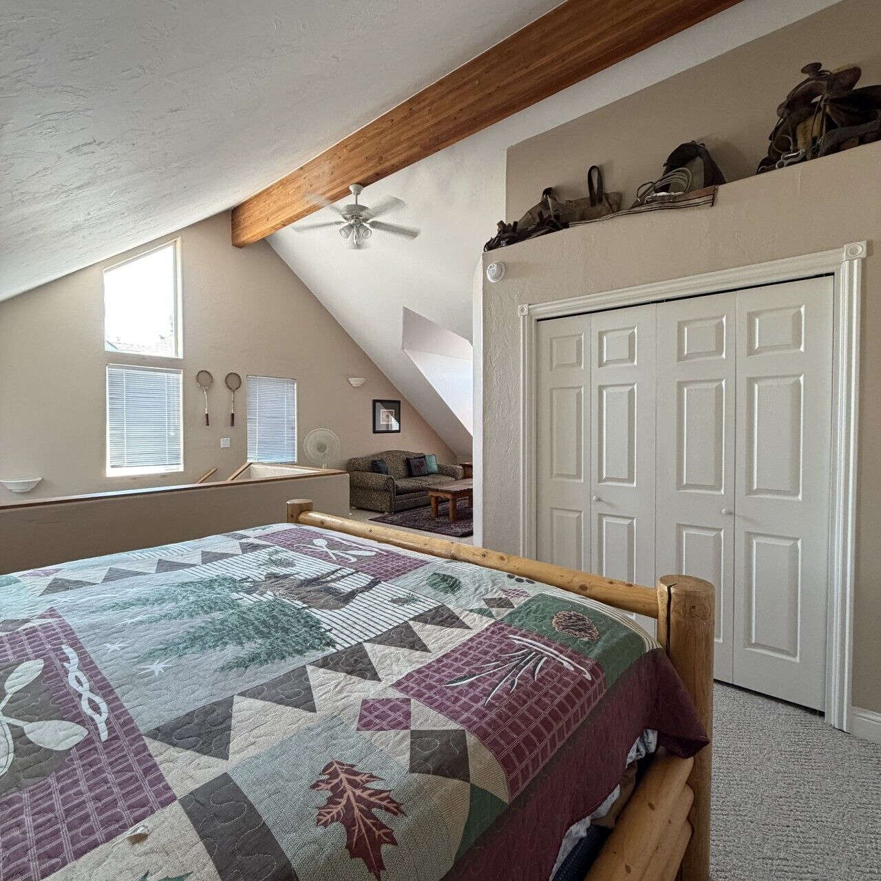 Ein Schlafzimmer mit schrägem Dach, einem Bett mit gemustertem Quilt, einem Deckenventilator, einem Fenster mit Jalousien, einer Tür und einer kleinen Sitzecke mit einer Couch.