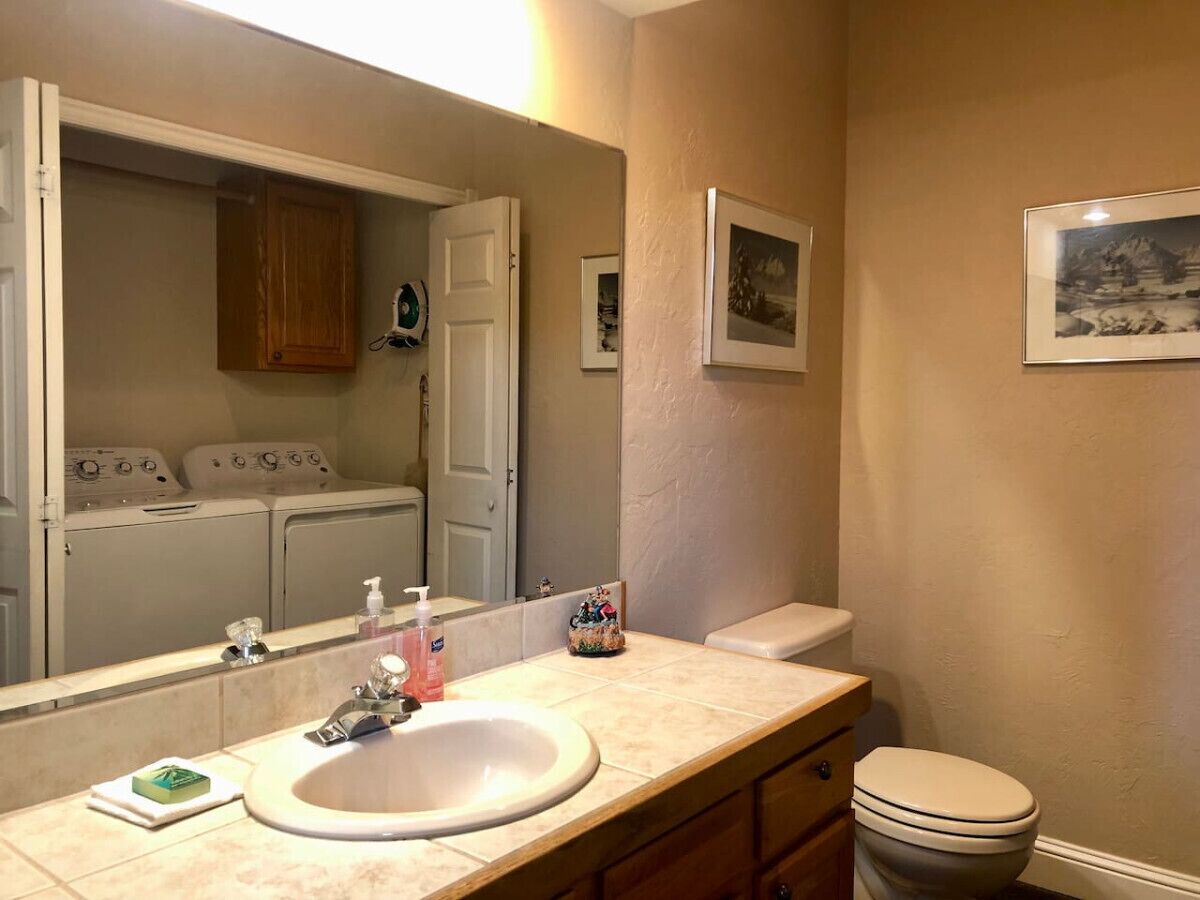 A bathroom with a sink, mirror, toilet, and a laundry area visible through an open door.