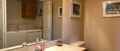 A bathroom with a sink, mirror, toilet, and a laundry area visible through an open door.