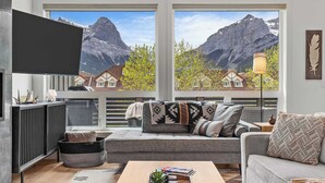 Una sala de estar moderna con un ventanal que ofrece vistas a la montaña, un televisor de pantalla plana en la pared y un rincón de descanso acogedor con un sofá y sillones.