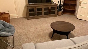 A living room with a grey sofa, a small round coffee table, a flat-screen TV on a cabinet, a potted plant, and a door. - Charming Brick Home in Seneca Falls Historic District! (Seneca Falls)