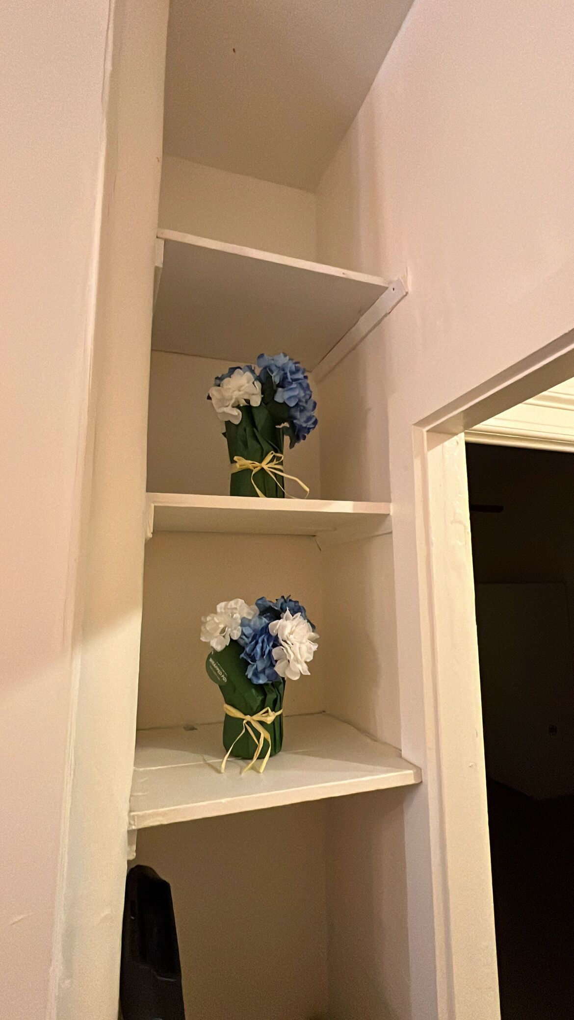 Eine Raumecke mit zwei weißen Regalen, auf jedem ein Blumenstrauß, gebunden mit einem gelben Band.