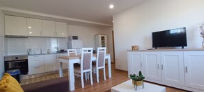 A modern kitchen with white cabinets, a dining area with a white table and chairs, a TV on a cabinet, and a small plant on a table. - Mondalva Apartamentos (Penacova)