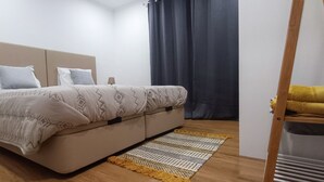 A bedroom with a bed, pillows, a bedside table, a lamp, a window with curtains, and a wooden floor. - Mondalva Apartamentos (Penacova)