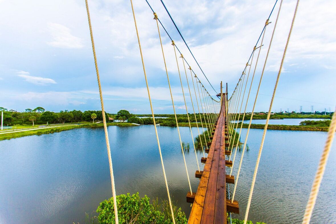 A wooden suspension bridge over a calm body of water with greenery on both sides.