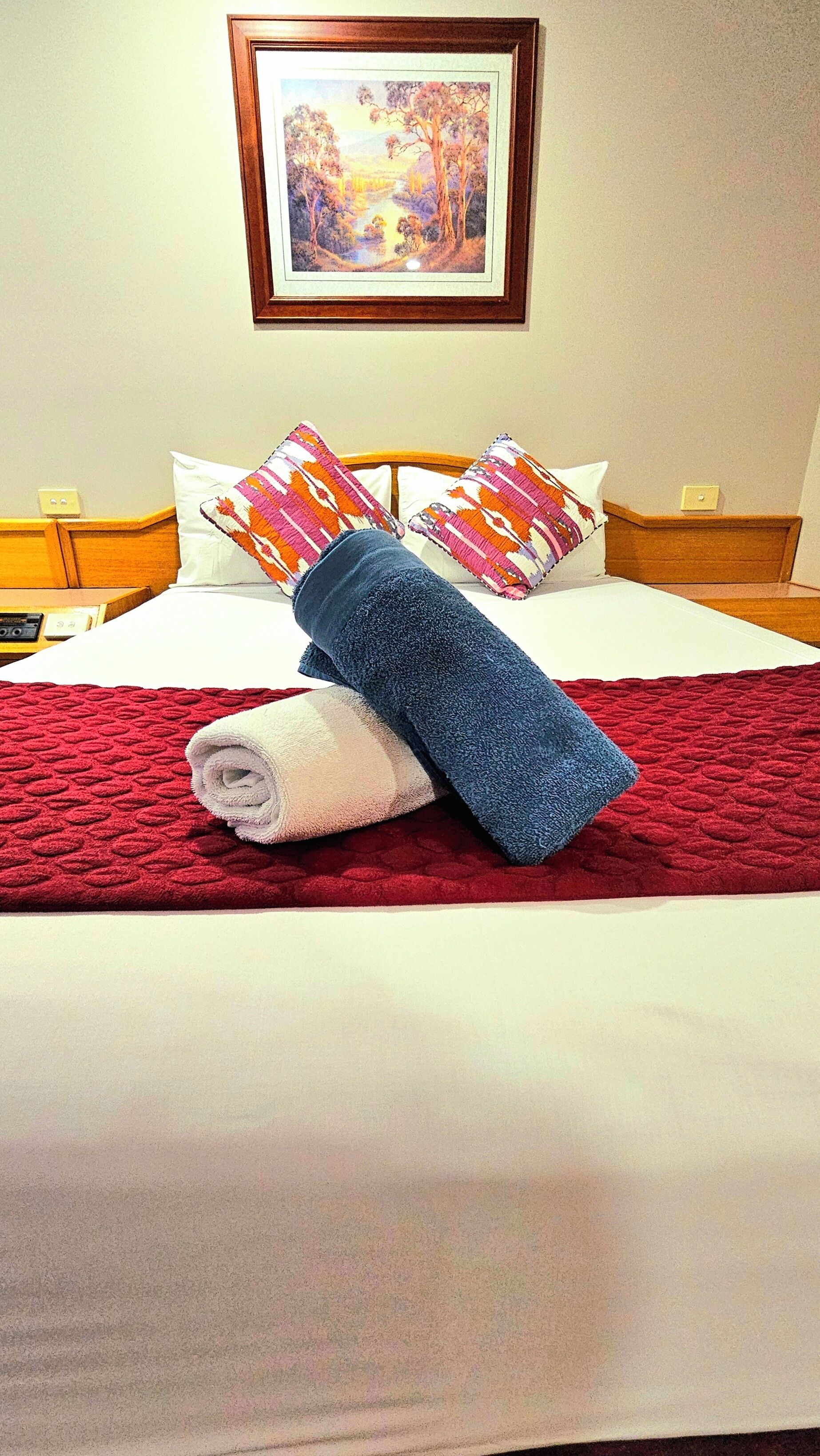 Una cama bien hecha con una manta roja, dos almohadas y toallas enrolladas.
