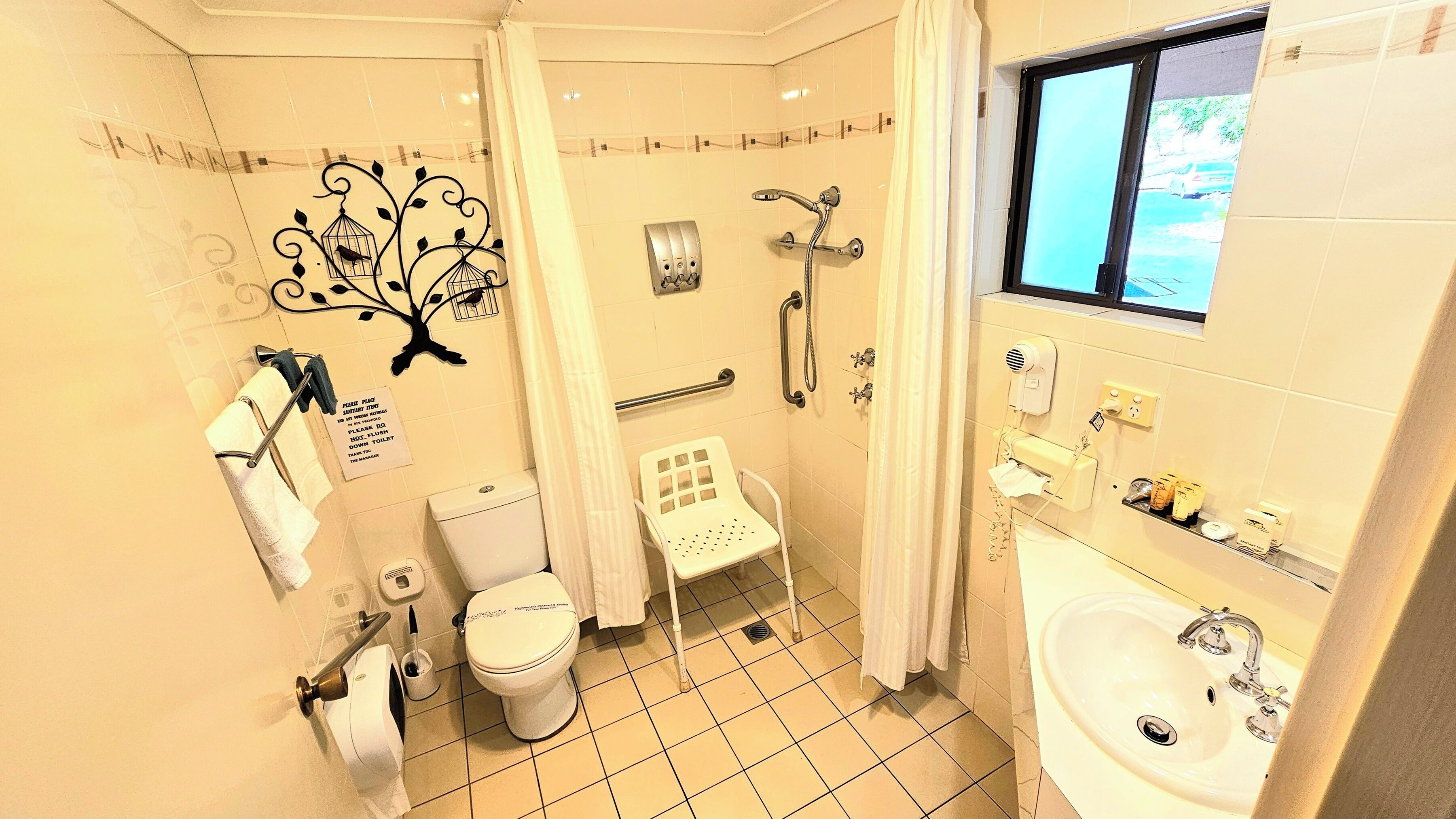 Un baño con inodoro, ducha y lavamanos. Hay un perchero, un secador de manos y un cartel en la pared.