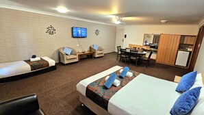 A hotel room with a bed, two armchairs, a small table, a desk with chairs, a television, and a kitchenette. - Hilltops Retreat Motor Inn (Young)