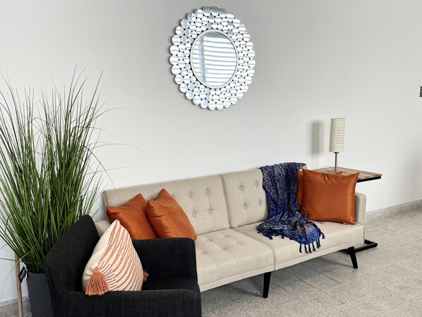Ein Wohnzimmer mit einem beigen Sofa, zwei orangen Kissen, einer blauen, gemusterten Decke und einer hohen Stehlampe.