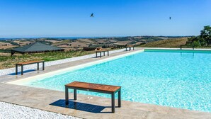 A swimming pool with a wooden table and benches, surrounded by a pebble border, overlooking a scenic landscape with hills and a clear sky. - Pet friendly apartment in Terre Roveresche (Terre Roveresche)