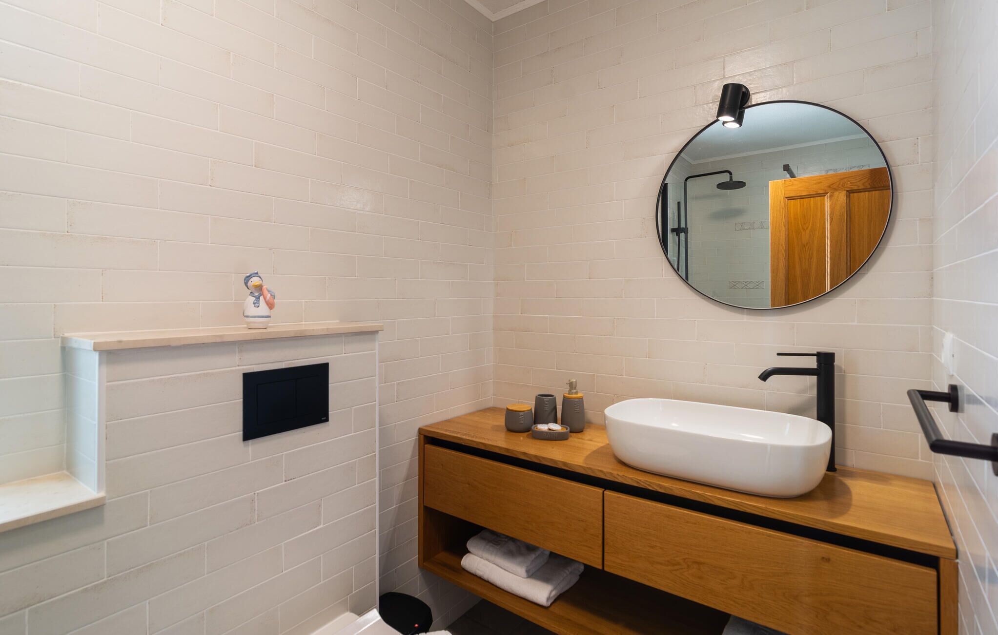 A modern bathroom with white brick walls, a wooden vanity, a white sink, and a round mirror.