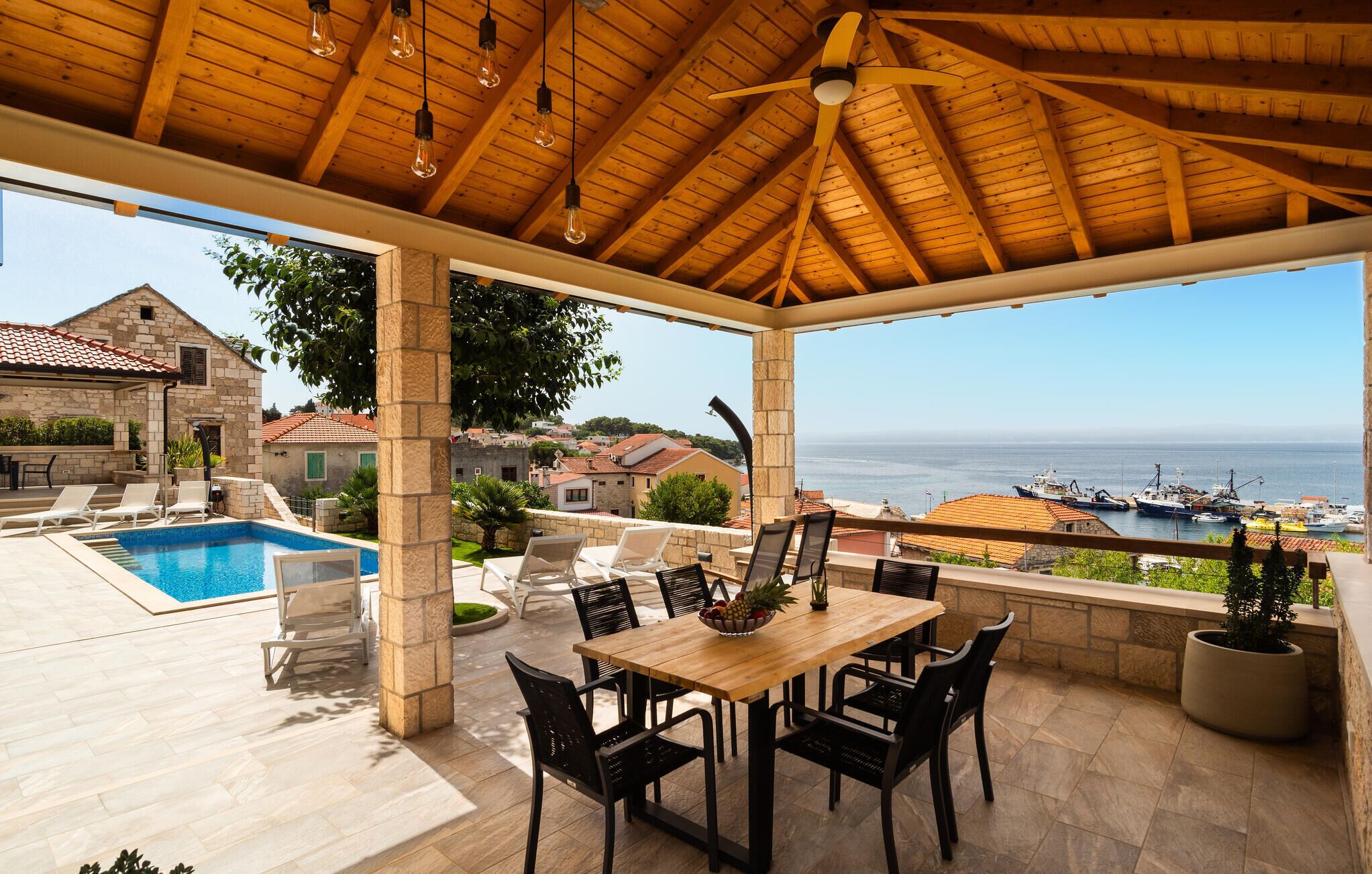 A covered outdoor area with a pool, dining table, and chairs, overlooking a harbor with boats.