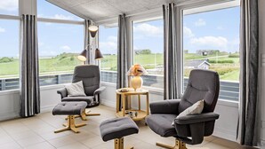 A modern room with two grey armchairs, a matching ottoman, and a small round table with a lamp. Large windows offer a view of a green landscape and a road. - Nice home in Lemvig with WiFi (Lemvig)
