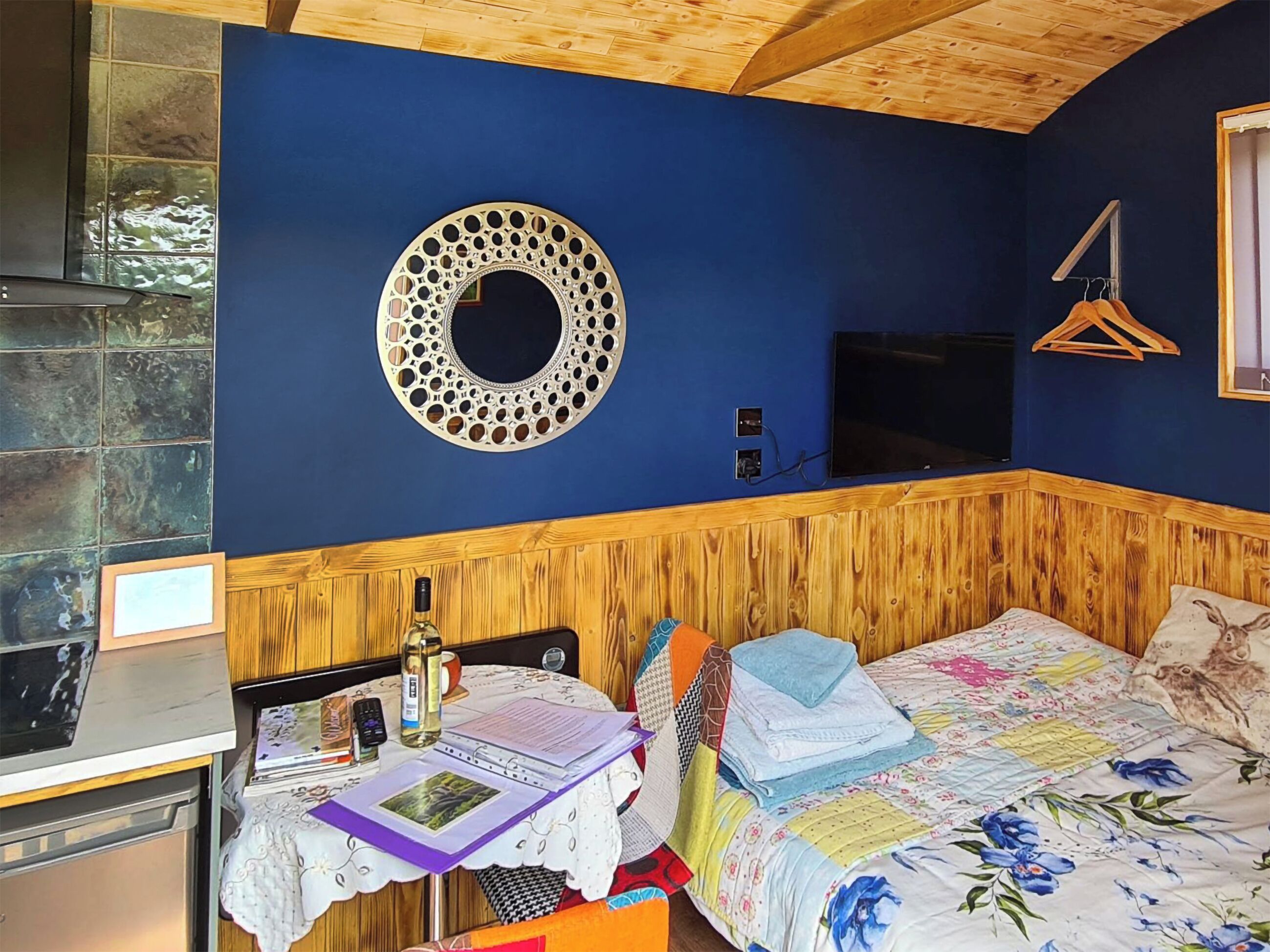A room with a wooden headboard, a circular wall decoration, a bed with floral bedding, a table with a laptop, and a hanging clothes rack.