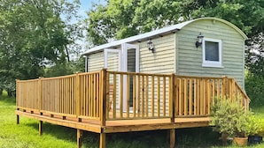 Eine kleine, eingeschossige Holzhütte mit einer Terrasse, umgeben von grünem Gras und Bäumen.