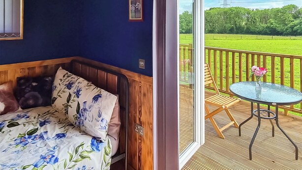 Ein Zimmer mit einem Bett, einer hölzernen Bank, einem kleinen Tisch mit einer Vase und Blick auf ein grünes Feld durch eine Glastür.