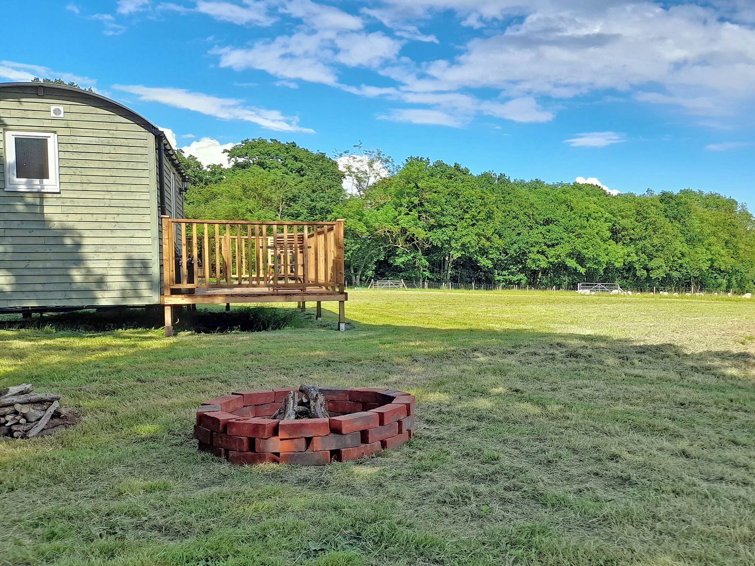 A wooden bench, a fire pit, and a trailer in a grassy area with trees in the background.