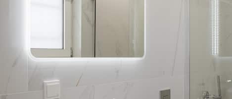 Un baño moderno con un lavamanos ovalado grande, una cabina de ducha de vidrio y un espejo rectangular con iluminación integrada.