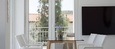 Une pièce avec une table en verre, des chaises blanches, un téléviseur fixé au mur et une vue sur les arbres et les bâtiments à l’extérieur.