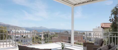 A balcony with wicker furniture, a small table with a plant, and a view of the sea and buildings.
