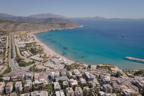 An aerial view of a coastal town with a beach, clear blue waters, and a mix of residential and commercial buildings. - Athenian Riviera Penthouse Apartment (Vari-Voula-Vouliagmeni)