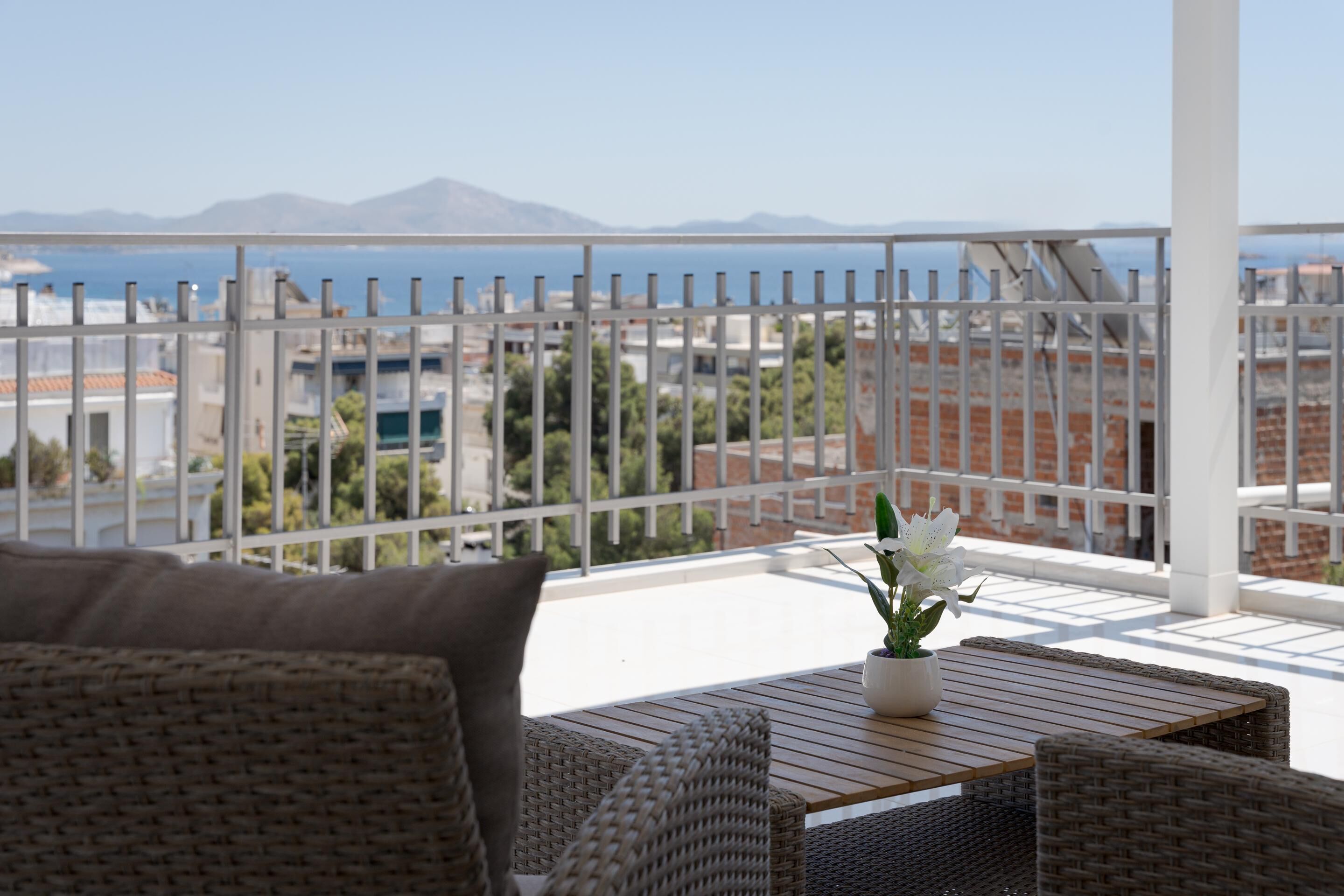 A balcony with wicker furniture, a potted plant, and a view of the sea and mountains.